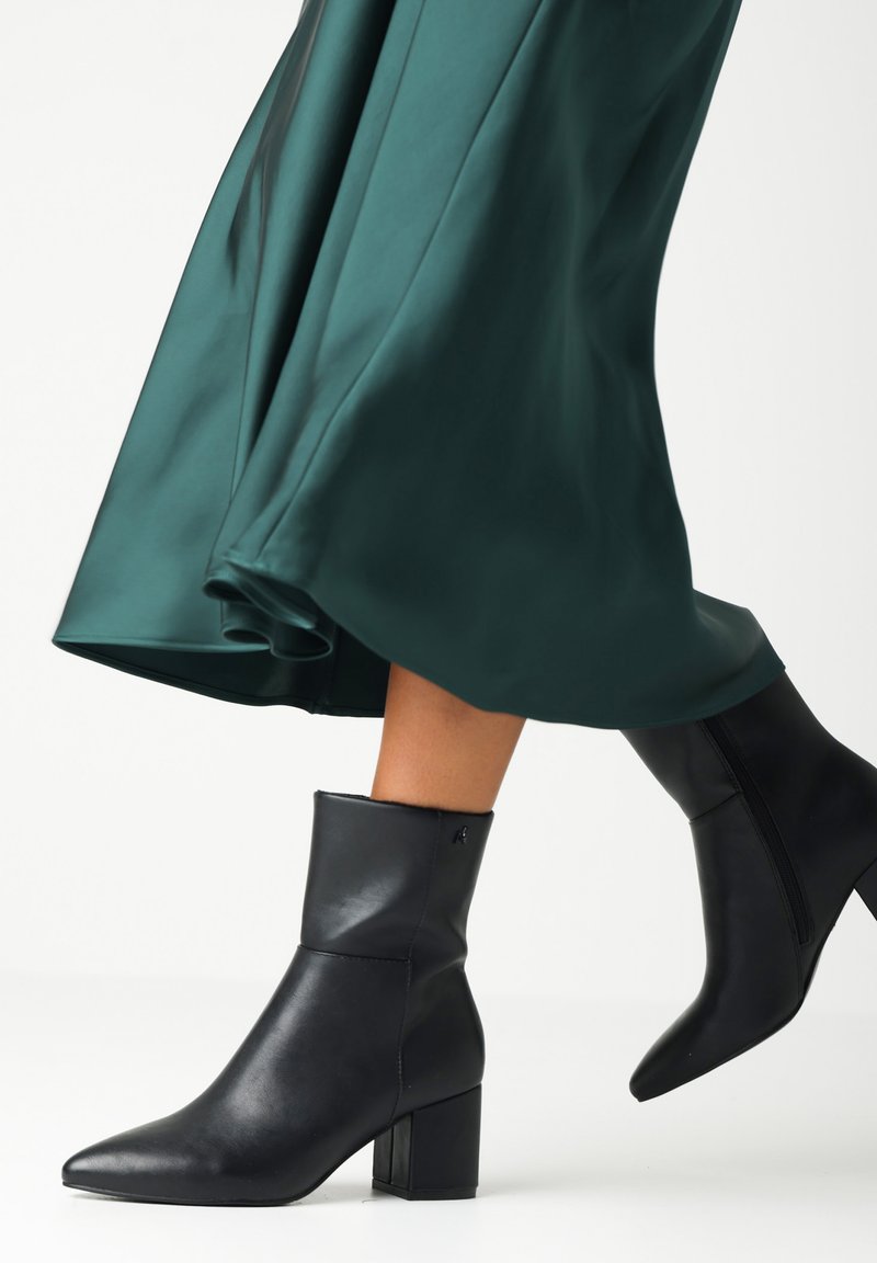 Botas de cuero negro hasta el tobillo con punta afilada y tacón cuadrado. La parte superior tiene una textura suave y hay un detalle de cremallera en un lado.