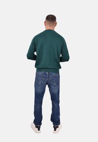 Grüner Strickpullover mit Rundhalsausschnitt, kombiniert mit blauen Jeans. Das Outfit wird durch weiße Sneakers mit schwarzen Akzenten vervollständigt.