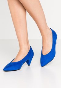 Chaussures à talons hauts en daim bleu avec un bout pointu et une découpe en forme de V à l'avant, reposant sur une surface blanche.