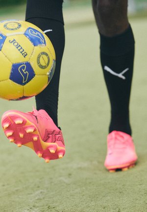 Rožinė futbolo batai su tekstūruota paviršiumi ir išsikišusiais kaiščiais, iškilęs logotipo detalė, padėta virš geltono kamuolio ant žalios žolės.