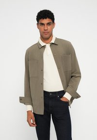 Oliivinvihreä napillinen takki, jossa on uurrettu kangas, kaksi rintataskua, päällä vaaleanbeige polo ja yhdistettynä tummansiniset farkut.