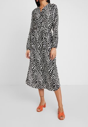 Mujer con vestido midi de manga larga con estampado de cebra en blanco y negro y cinturón anudado, combinado con sandalias de tacón naranja.