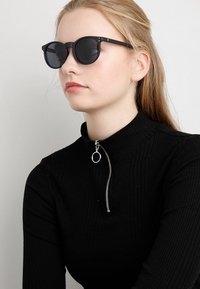 Ochelari de soare negri cu lentile rotunde și un cadru metalic negru elegant, purtați cu un top negru cu guler înalt din material texturat, având un detaliu cu fermoar argintiu.