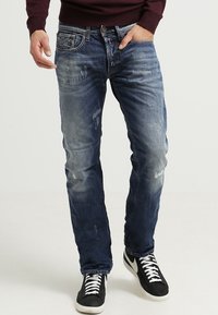Jeans de mezclilla en azul oscuro con un acabado desgastado, diseño slim fit, cinco bolsillos y patrones de desvanecimiento notables. Combinados con zapatillas negras.