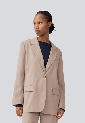 Blazer beige ajusté avec revers crantés, fermeture à un bouton, deux poches avant et tissu texturé, associé à un haut sombre.
