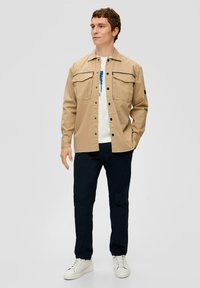  lichtbruine knoopjack met twee borstzakken en zwarte accenten, gedragen over een wit T-shirt, gecombineerd met donkere broeken en witte sneakers.