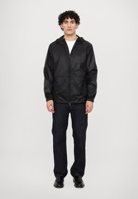 Veste légère noire avec capuche, fermeture éclair à l'avant et poignets élastiques. Portée par-dessus une chemise blanche, associée à un jean foncé et des chaussures noires.