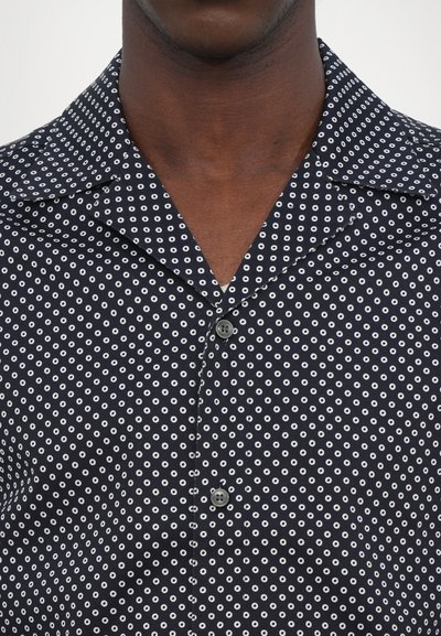 Chemise bleu marine à manches courtes avec un motif circulaire blanc, col boutonné, et un seul bouton gris à l'avant. Texture de tissu lisse.