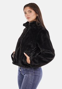 Giacca in pelliccia sintetica nera con colletto alto, zip frontale e tasche laterali, indossata da un modello con jeans blu.