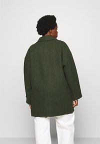 Oversized, dark green coat made of textured fabric with a collared neckline. Features dropped shoulders and a straight cut.