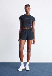 Czarny krótki t-shirt z logo "Nike", w połączeniu z czarnymi szortami. Model nosi białe tenisówki i skarpetki na niebieskim dywanie.