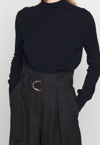 Pull à col roulé en tricot bleu marine associé à un pantalon noir taille haute avec une large ceinture et une boucle circulaire en argent.