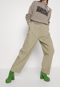 Grijze gebreide trui met zwarte opschriften, gecombineerd met beige hoog getailleerde plooibroek en groene platformhakken met een gestructureerde oppervlakte.