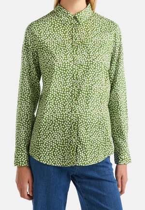 Frau trägt ein grünes, langärmliges Hemd mit weißen Punkten und blaue Jeans, steht vor hellem Hintergrund.