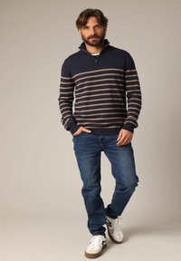 Pull marin avec des rayures brunes, col zippé, associé à un jean bleu et des chaussures blanches. Texture douce et coupe décontractée. Style décontracté.
