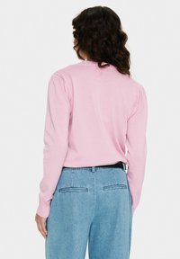 Femme aux cheveux foncés et bouclés portant un pull rose à manches longues et un jean taille haute bleu clair, vue de dos sur un fond uni.