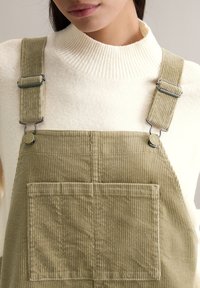 Olijfgroene ribfluwelen tuinbroek met verstelbare metalen clips en zakdetail, gedragen over een crèmekleurige geribbelde trui. Close-up van de stoftextuur.