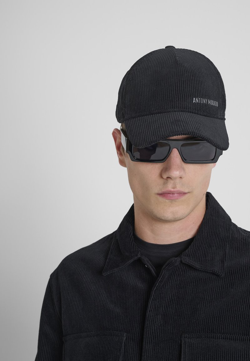 Crna kapica od kordura s natpisom "ANTONY MORATO", u kombinaciji s crnim pravokutnim naočalama i crnom kordura jaknom.