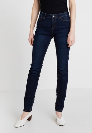 Personne portant un jean slim bleu foncé et des chaussures blanches à talons, debout devant un fond clair uni.