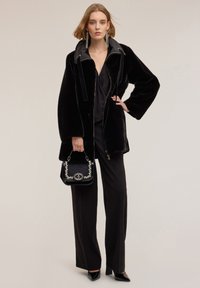 Czarna kurtka z szerokimi rękawami i zamkiem z przodu, wykonana z sztucznego futra, zestawiona z eleganckim czarnym topem i spodniami z wysokim stanem. Uzupełniona ozdobną torebką.