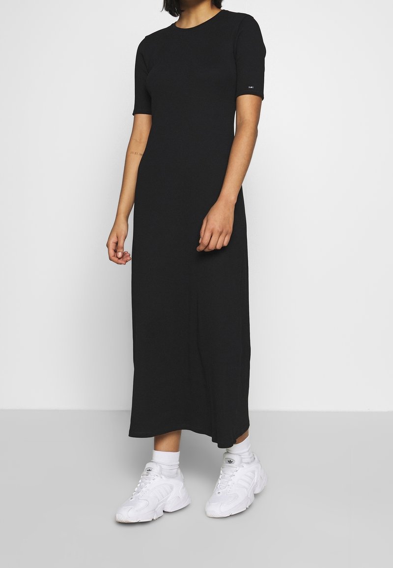 Femme portant une robe midi noire à manches courtes et des baskets blanches, debout devant un fond gris clair uni.