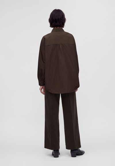 Personne aux cheveux courts et foncés portant une chemise ample à manches longues brun foncé et un pantalon large assorti, debout de dos sur un fond uni.