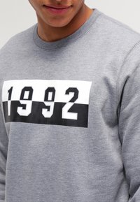 Grå sweatshirt med rund halsringning och en djärv grafisk design i vitt och svart som visar texten "1992" över bröstet. Mjuk textur.
