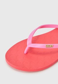 Rosa flip-flop med en struktureret rød gummisål, der har en rosa tætsiddende rem og en guldfarvet logoakcent på remmen.