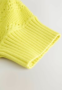 Heller gelber Strickpulloverärmel mit gerippter Textur; verfügt über ein grobes Muster und ordentliche Stichdetails am Bündchen.