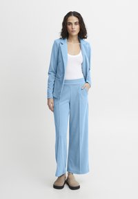 Lichtblauw stoffen pak met een op maat gemaakte blazer en brede broeken, met een gladde textuur en losse pasvorm. Witte tanktop eronder, casual schoeisel.