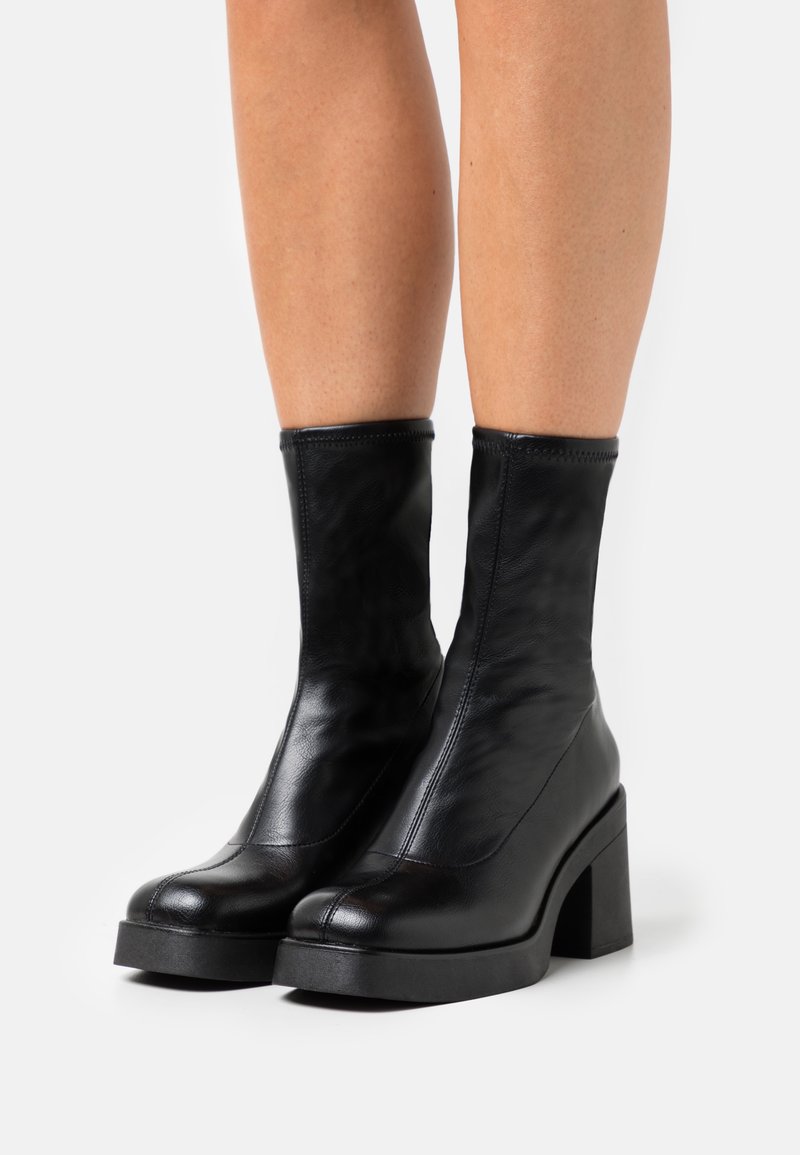 Bottes en cuir noir à talons carrés portées sur des jambes nues, sur fond blanc.