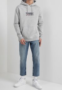 Sudadera gris con bolsillo frontal, con el logo "TOMMY JEANS" en azul marino y rojo. Combinada con jeans de color azul claro y zapatillas blancas.