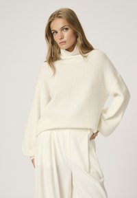Pull en maille blanche avec un col montant, coupe oversize et manches larges ; associé à un pantalon beige. Texture douce, design minimaliste.