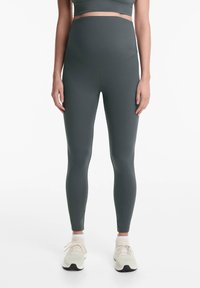 Sötétzöld, magas derékú leggings, rugalmas anyagból készült. Sima textúrájú, testhezálló kivitel, világos sportcipőkkel párosítva.