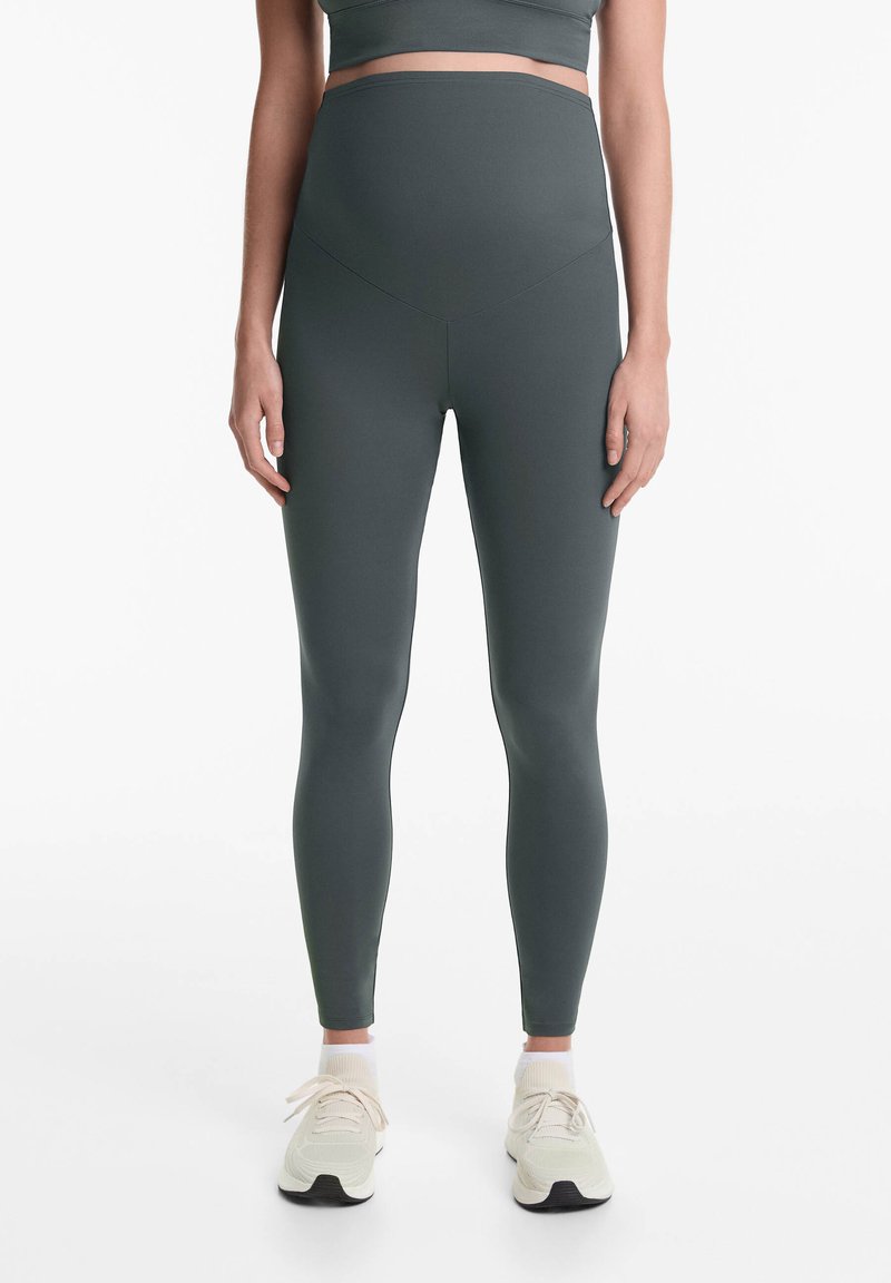 Sötétzöld, magas derékú leggings, rugalmas anyagból készült. Sima textúrájú, testhezálló kivitel, világos sportcipőkkel párosítva.