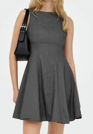 Kvinde iført ærmeløs, mørkegrå, vid nederdel kjole, bærende en lille sort skuldertaske på venstre skulder mod en ensfarvet baggrund.