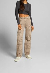 Pantalones cargo beige con múltiples bolsillos laterales, un corte holgado y una resistente mezcla de algodón. Combinados con un top corto de canalé oscuro y zapatillas ligeras.