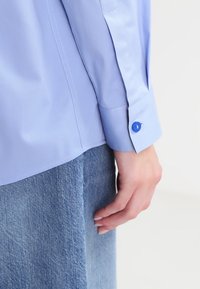 Camicia azzurra chiara realizzata in tessuto liscio, con una vestibilità su misura, maniche lunghe e chiusura a bottoni con un bottone blu a contrasto.