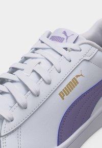 Λευκό αθλητικό παπούτσι Puma με λιλά ρίγα, χρυσό λογότυπο Puma στο πλάι και λευκά κορδόνια δεμένα στην κορυφή.