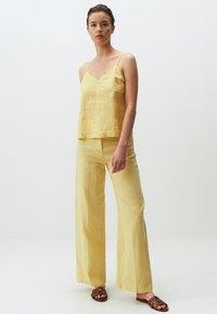 Geel linnen tanktop met dunne banden en een V-hals, gecombineerd met wijde gele linnen broeken. Bruine sandalen met banden maken de outfit af.