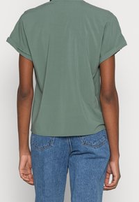 Persona che indossa una camicia verde a maniche corte e jeans in denim blu, vista di spalle contro uno sfondo neutro.