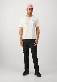 Bílé tričko s krátkým rukávem a kruhovým logem, černé cargo kalhoty s bočními kapsami a růžová čepice s kulatým emblémem. Černé boty.