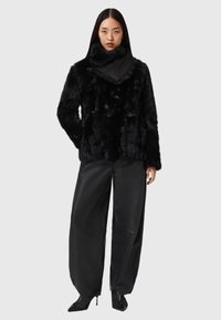 Giacca in pelliccia ecologica nera con collo alto, abbinata a pantaloni larghi in pelle nera e stivali neri con tacco a punta, su sfondo neutro.
