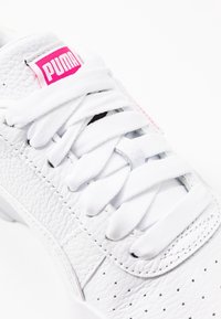 White sneaker with textured leather upper, white laces, and pink logo tag. Features perforations and stitching details on the side.
