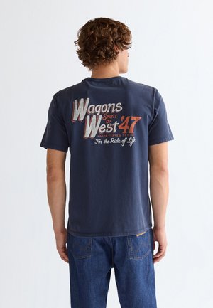 Νέος με σγουρά μαλλιά, φορώντας μπλε σκούρο μπλουζάκι και τζιν, με την πλάτη προς την κάμερα· στο μπλουζάκι αναγράφεται "Wagons Spirit of West '47."