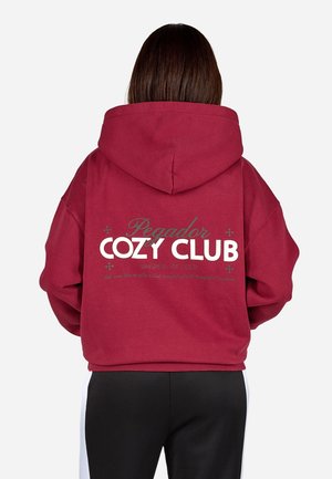 Persona con cabello largo y castaño que lleva una sudadera con capucha roja con el texto "Pegador Cozy Club" en la espalda, combinada con pantalones negros y blancos.