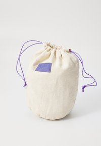 Cotton drawstring bag in cream, featuring purple cord and a rectangular leather patch. Rounded shape with gathered top and smooth texture.