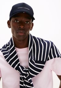 Gorra de béisbol navy con el logo de "TOMMY HILFIGER"; camiseta rosa; suéter a rayas navy y blancas atado al cuello, creando un look en capas.