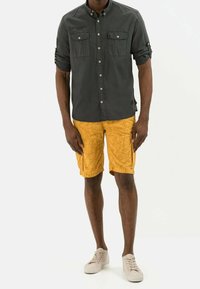 Chemise grise à manches retroussées, associée à un short jaune avec un motif de feuilles. Des chaussures claires complètent la tenue.