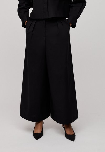 Black wide-leg trousers made of smooth fabric feature pleats, side pockets, and a relaxed fit, paired with black pointed-toe heels.